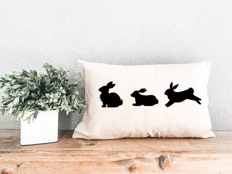 May include: A white pillow with three black bunny silhouettes. The bunnies are in different poses, one is sitting, one is standing, and one is hopping.
