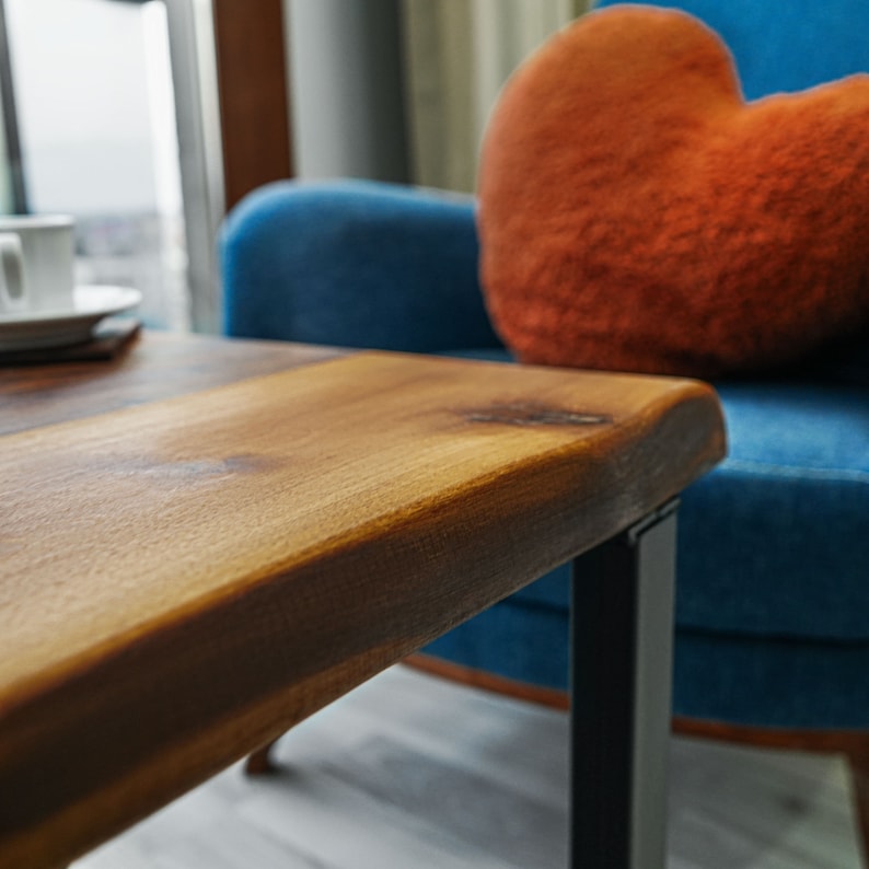 Op de afbeelding: Een close-up van een houten salontafel met een live edge blad en zwarte metalen poten. De tafel staat voor een blauwe fauteuil met een oranje hartvormig kussen.