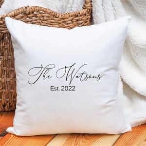 May include: A white square pillow with the text "The Watsons" and "Est. 2022" in black script. The pillow is resting on a wooden surface with a woven basket and a white blanket in the background.