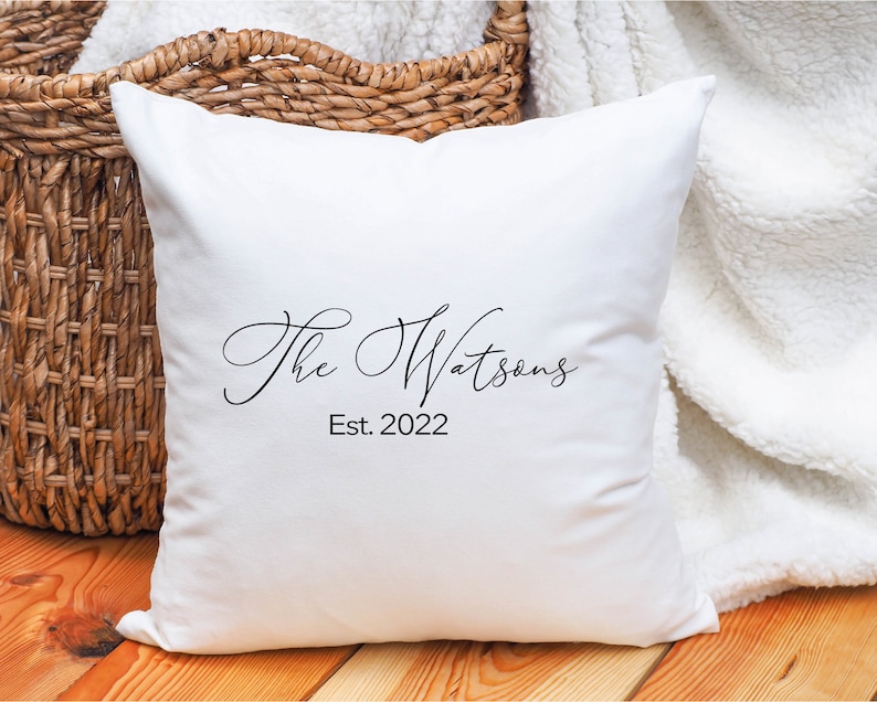 May include: A white square pillow with the text "The Watsons" and "Est. 2022" in black script. The pillow is resting on a wooden surface with a woven basket and a white blanket in the background.