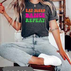 May include: Heather dark grey t-shirt with the text "Eat Sleep Dance Repeat" in colourful, retro-style lettering.