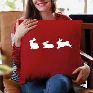 May include: A red throw pillow with a white silhouette of three rabbits. The rabbit on the right is hopping.