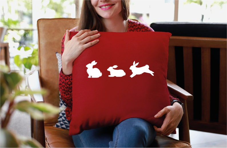May include: A red throw pillow with a white silhouette of three rabbits. The rabbit on the right is hopping.
