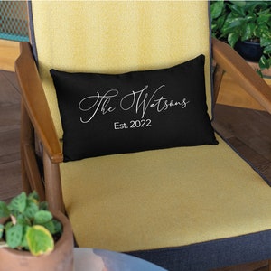 May include: A black rectangular pillow with the text "The Watsons" in a white script font and "Est. 2022" below it. The pillow is on a yellow chair with wooden arms.