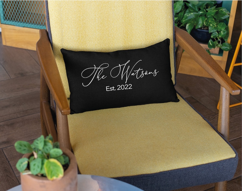 May include: A black rectangular pillow with the text "The Watsons" in a white script font and "Est. 2022" below it. The pillow is on a yellow chair with wooden arms.