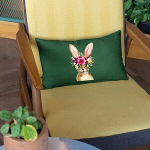 May include: A green throw pillow with a brown bunny wearing a flower crown. The pillow is on a yellow chair with a wooden frame.