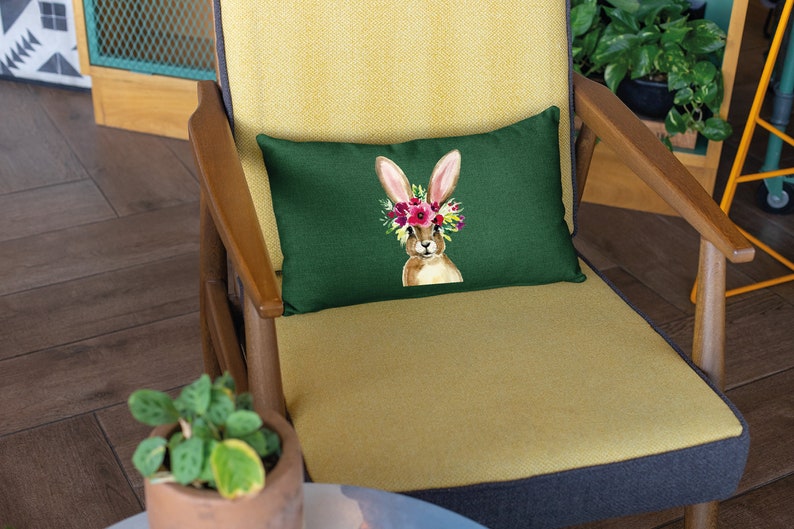 May include: A green throw pillow with a brown bunny wearing a flower crown. The pillow is on a yellow chair with a wooden frame.