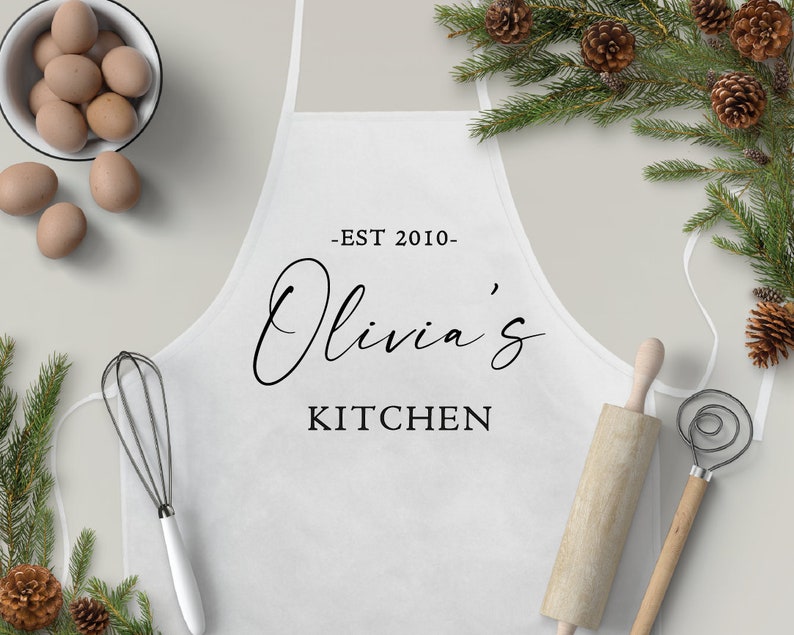 May include: White kitchen apron with the text "EST 2010 - Olivia's Kitchen" printed in black.