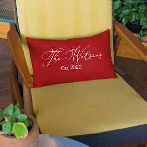 May include: A rectangular red decorative pillow with the text "The Watsons Est. 2022" in white script. The pillow is placed on a yellow chair with wooden arms.