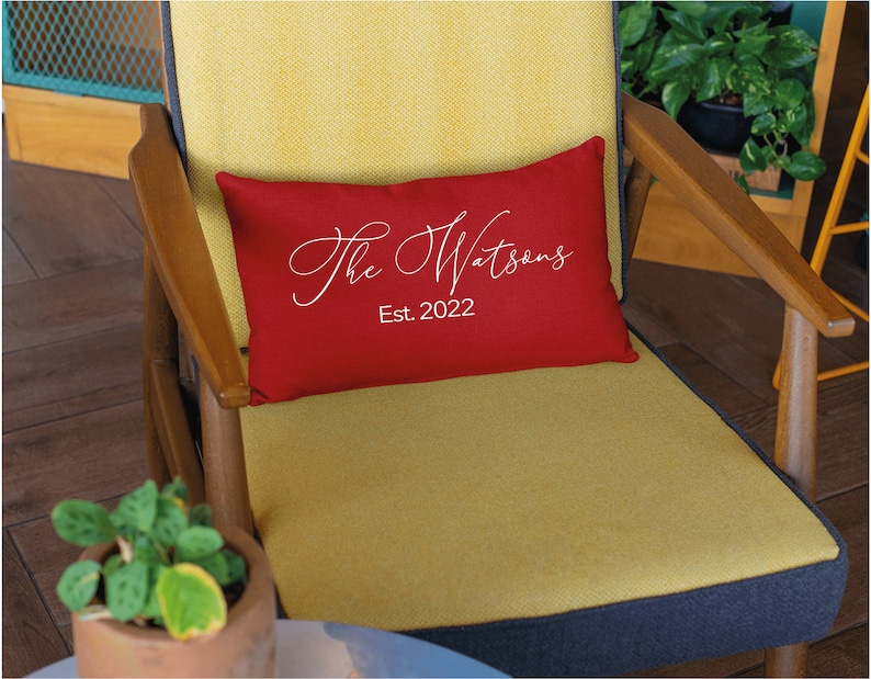 May include: A rectangular red decorative pillow with the text "The Watsons Est. 2022" in white script. The pillow is placed on a yellow chair with wooden arms.