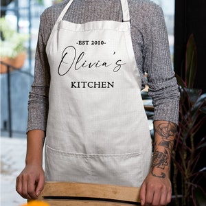 May include: White kitchen apron with black text that reads "Est 2010 - Olivia's Kitchen".
