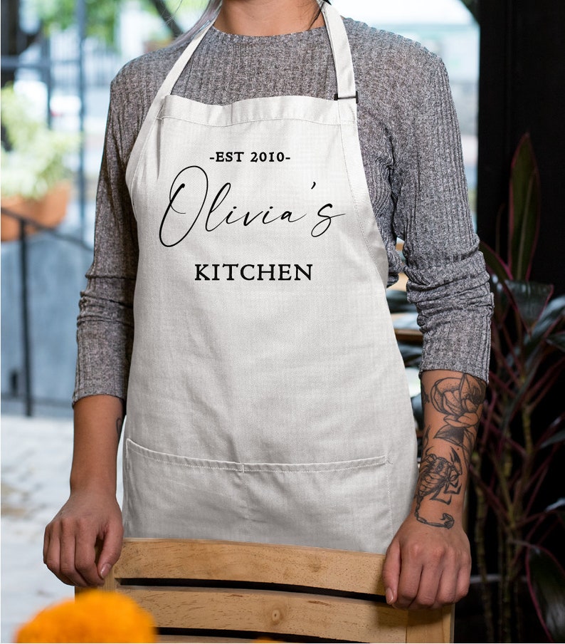 May include: White kitchen apron with black text that reads "Est 2010 - Olivia's Kitchen".