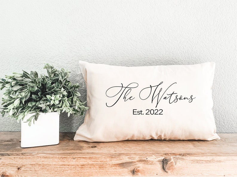 May include: Cream-colored rectangular pillow with the text "The Watsons" in a cursive font and "Est. 2022" below it. The pillow is on a wooden surface, with a white square vase holding green plants to the left.