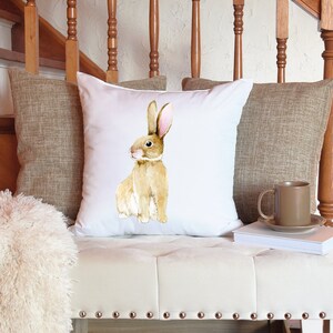May include: A white cushion with a watercolour illustration of a brown bunny rabbit. The cushion is sitting on a light brown sofa with a brown throw blanket and a brown mug on a book.