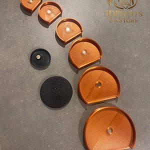 May include: Orange 3D-printed semi-circular objects with a central hole and a small magnet, plus a black circular object and a black disc. The items are arranged on a grey surface. The text "3DPRINTS UKSTORE" is visible in the upper right corner.