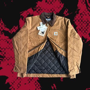 Peut inclure: Une veste Carhartt beige avec un col en velours côtelé marron et une doublure matelassée noire. La veste présente une fermeture éclair frontale, une poche poitrine zippée et un écusson Carhartt sur la poitrine. Plusieurs étiquettes sont attachées.