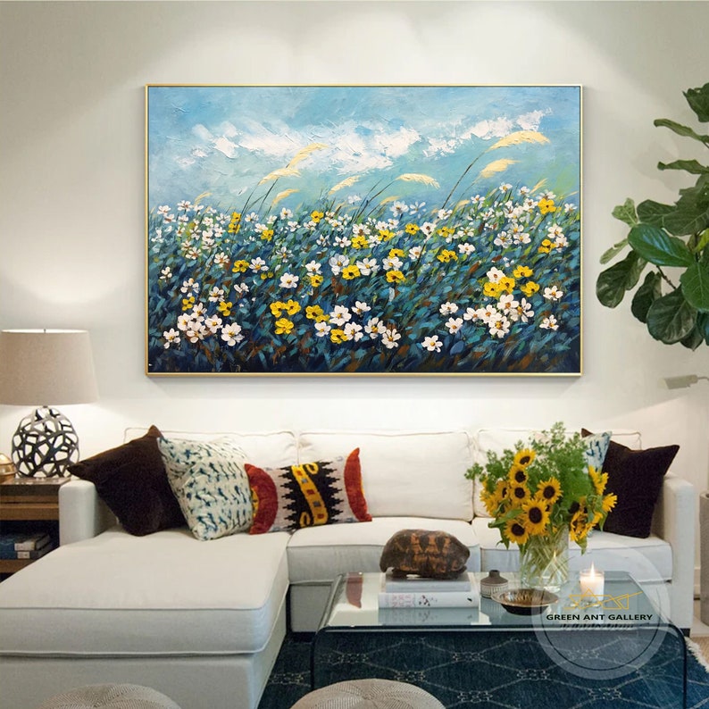 K&ouml;nnte beinhalten: Ein Gem&auml;lde, das ein Feld mit wei&szlig;en und gelben Blumen mit blauem Himmel und wei&szlig;en Wolken im Hintergrund zeigt. Das Gem&auml;lde ist in Gold gerahmt und h&auml;ngt an einer wei&szlig;en Wand &uuml;ber einem wei&szlig;en Sofa.