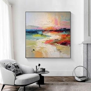 May include: Abstract landscape painting with a black frame. The artwork features a textured, colorful composition with hues of white, blue, pink, orange, red, and yellow. The painting is displayed above a gray armchair and a small black side table.
