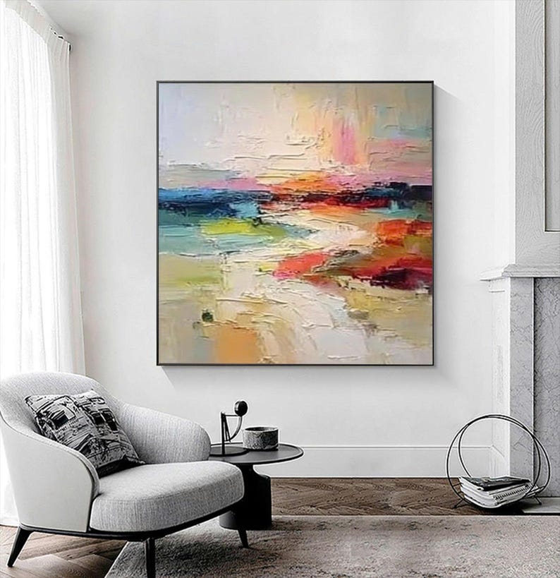 May include: Abstract landscape painting with a black frame. The artwork features a textured, colorful composition with hues of white, blue, pink, orange, red, and yellow. The painting is displayed above a gray armchair and a small black side table.