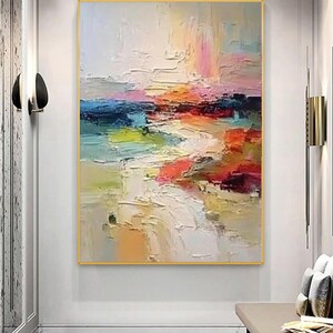 May include: Abstract landscape painting with a gold frame. The artwork features a textured, colorful composition with strokes of white, blue, pink, orange, and green. The painting is displayed on a white wall.