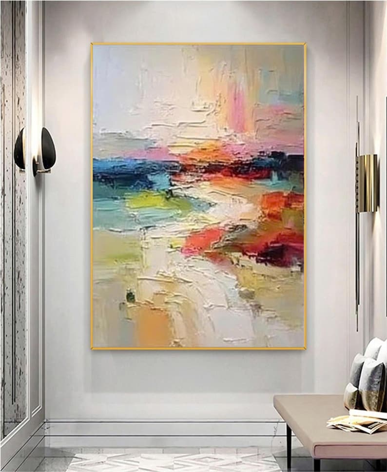 May include: Abstract landscape painting with a gold frame. The artwork features a textured, colorful composition with strokes of white, blue, pink, orange, and green. The painting is displayed on a white wall.