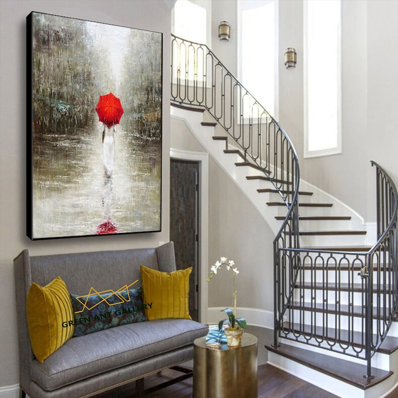 May include: A painting of a person walking in the rain with a red umbrella. The painting is framed and hanging on a wall in a home interior setting.