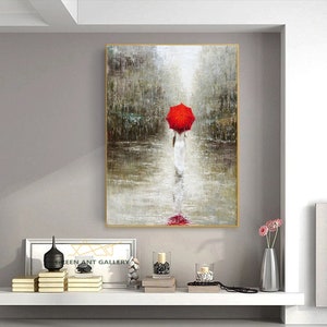 May include: A painting of a woman walking in the rain with a red umbrella. The painting is in a gold frame and is hanging on a wall. The woman is wearing a white dress and is walking away from the viewer. The background is a blurry image of a city street.