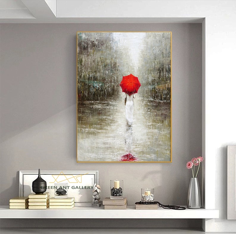 May include: A painting of a woman walking in the rain with a red umbrella. The painting is in a gold frame and is hanging on a wall. The woman is wearing a white dress and is walking away from the viewer. The background is a blurry image of a city street.