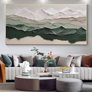 May include: Abstract landscape painting with white, beige, and green colors. The painting has a textured, layered look, giving the impression of mountains or hills. The painting is framed in a gold frame and is hanging on a white wall.