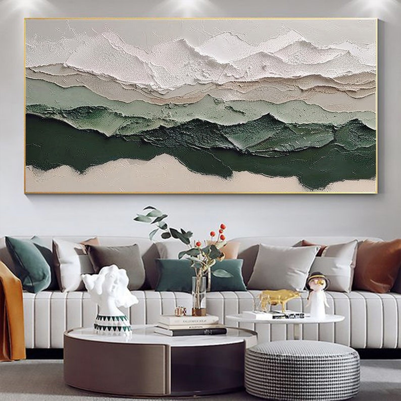 May include: Abstract landscape painting with white, beige, and green colors. The painting has a textured, layered look, giving the impression of mountains or hills. The painting is framed in a gold frame and is hanging on a white wall.