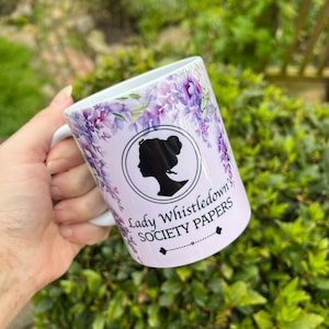 May include: White ceramic mug with a purple floral design and a black silhouette of a woman's head. The text "Lady Whistledown's Society Papers" is printed on the mug.