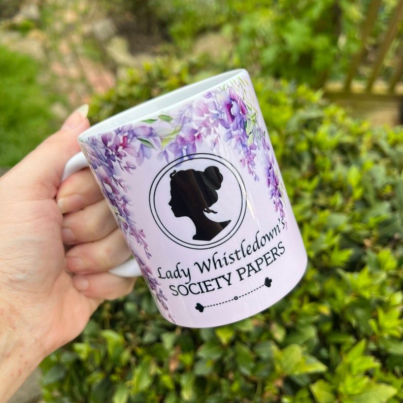 May include: White ceramic mug with a purple floral design and a black silhouette of a woman's head. The text "Lady Whistledown's Society Papers" is printed on the mug.