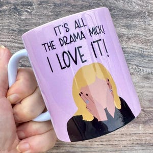 May include: A pink ceramic mug with a white handle. The mug features a black illustration of a woman with blonde hair covering her face with her hands. The text on the mug reads "It's all the drama Mick! I love it!"