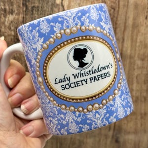 Puede incluir: Una taza de cer&aacute;mica blanca con un dise&ntilde;o floral azul y el texto "Lady Whistledown's SOCIETY PAPERS". La taza presenta un &oacute;valo con borde dorado con la silueta de una mujer. El asa es blanca.