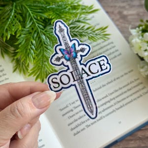 May include: A holographic sticker featuring a sword with a butterfly design and the word "SOLACE" in a white font.