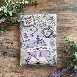 May include: A clear phone case with a light blue background, featuring book-themed illustrations and text. The design includes a sword, open book, keys, and the words "Forged by Fiction" and "Certified Book Girly."