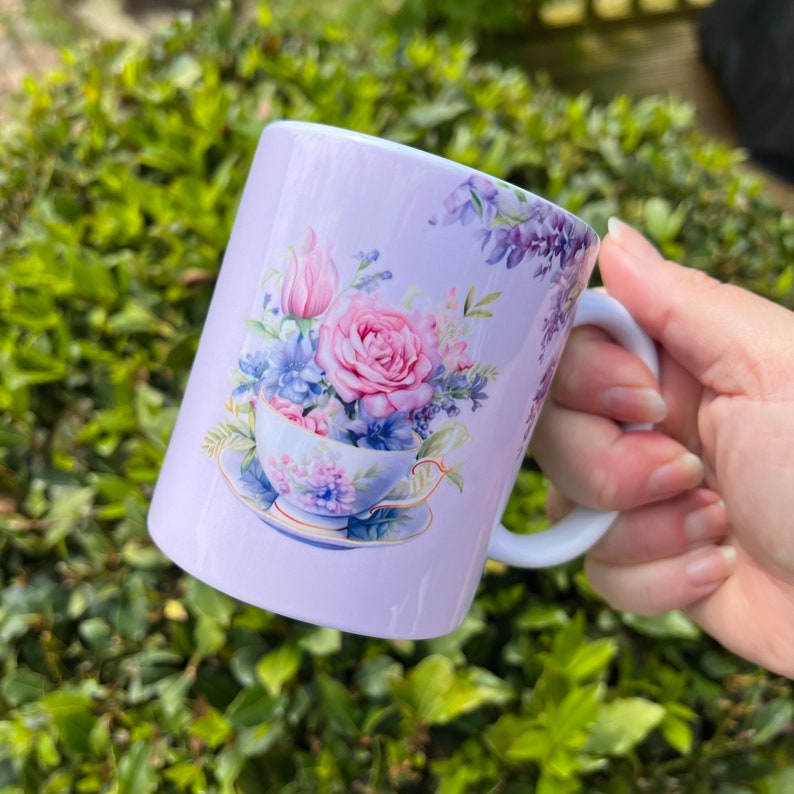 May include: A lavender mug with a floral design featuring a teacup filled with pink, purple, and white flowers. The flowers are surrounded by green leaves and vines.