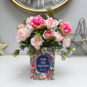 May include: A floral arrangement of pink and pale pink roses and foliage in a decorative vase. The vase is patterned with flowers, a peacock illustration, and the text "Pride and Prejudice" by Jane Austen.