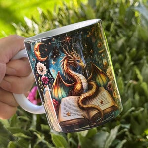 May include: White ceramic mug with a fantasy illustration. It depicts a golden dragon, an open book, a crescent moon, stars, and flowers. The mug has a white handle and a glossy finish, ideal for tea or coffee.