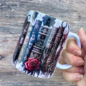 May include: White ceramic mug with a design of stacked, ornate books in purple, black, and red. A red rose is at the base, and the text "I Prefer the Villain" is displayed. The mug has a white handle.