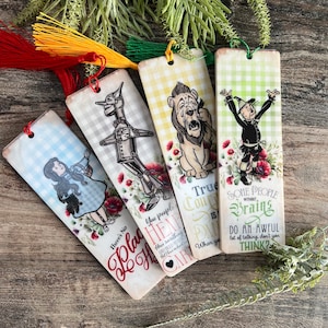 May include: Four rectangular bookmarks with tassel details. Each bookmark features a character from The Wizard of Oz, including Dorothy, the Tin Man, the Cowardly Lion, and the Scarecrow, with quotes and floral designs. The bookmarks have a vintage, distressed appearance.