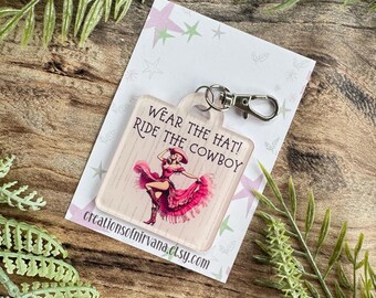 May include: A white square keychain with a pink and white image of a woman wearing a hat and a dress. The text on the keychain reads "Wear the hat! Ride the cowboy!"