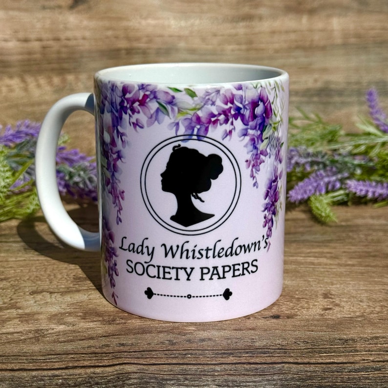 May include: White ceramic mug with a purple floral design and a silhouette of a woman's head. The text "Lady Whistledown's Society Papers" is printed on the mug.