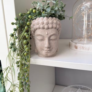 May include: A beige Buddha head planter filled with greenery and trailing vines. The planter has a serene expression and detailed hair. The trailing vines have small, round, green beads. The planter is on a white shelf.