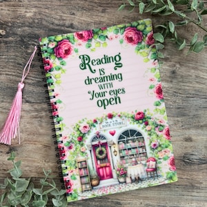 May include: Spiral-bound notebook with a pink cover featuring a floral design and the text "Reading is dreaming with your eyes open." The cover also depicts a bookstore scene with a pink door and bookshelves. A pink tassel bookmark is attached.