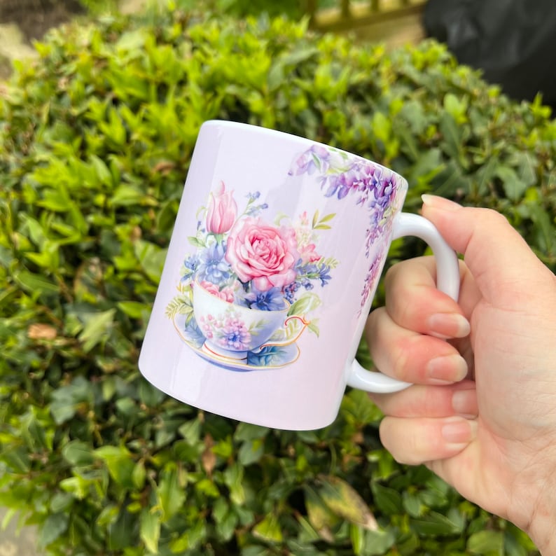 May include: A white ceramic mug with a floral design featuring a teacup filled with pink and purple flowers. The mug has a white handle and is set against a green background.