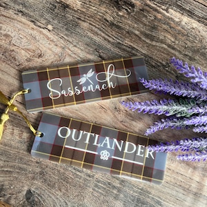 May include: Two grey and brown plaid bookmarks with gold tassels. The top bookmark has the word "Sassenach" written in white with a dragonfly design. The bottom bookmark has the word "OUTLANDER" written in white with a knot design.