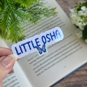 May include: A clear sticker with a glittery background and a blue butterfly design. The sticker says "LITTLE OSHA" in blue letters.
