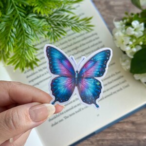 May include: A blue and purple butterfly sticker with glitter accents. The butterfly has a white background and is on a book.