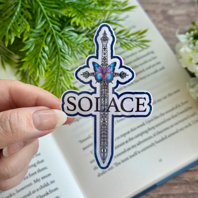May include: A holographic sticker with a sword and butterfly design. The sword is silver with a black handle and the butterfly is blue and purple. The word "SOLACE" is written in black letters on the sticker.